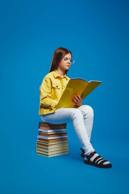 Parlak bir stüdyoda, gözlüklü meraklı bir genç kitap yığınının üzerinde oturuyor, okumaya dalmış, öğrenme tutkusunu somutlaştırıyor. Eğitim materyalleri ve kampanyalar için ideal.