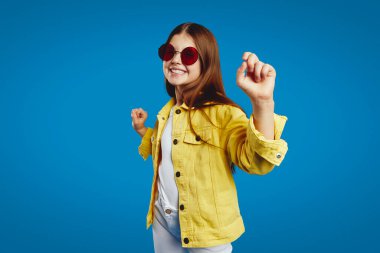 Canlı sarı bir ceket giyen neşeli bir çocuk ve modaya uygun güneş gözlükleri, mutluluk ve enerjiyi ifade eden parlak mavi bir arka plana karşı coşkulu bir şekilde dans ediyor. Reklam ve marka kampanyaları için ideal..