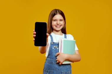 Okul kitaplarını tutarken akıllı telefon görüntüsü sergileyen çekici küçük kız neşe ve merak saçıyor. Eğitimsel promosyonlar ve teknolojiyle ilgili reklamlar için ideal.