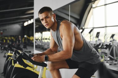 Spor giyimli, spor kıyafetli, spor salonunda spor bisikletle egzersiz yapan bir adam. Sporcu erkek egzersiz bisikleti eğitimi alıyor ve kameraya bakıyor. Genç adam modern spor salonundaki bir sınıfta çalışıyor..