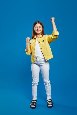 Parlak sarı ceketli mutlu genç, mavi arka planda izole edilmiş, heyecan ve başarısını ifade eden yumruklarını kaldırıyor. Olumlu ve gençlere yönelik pazarlama için ideal.