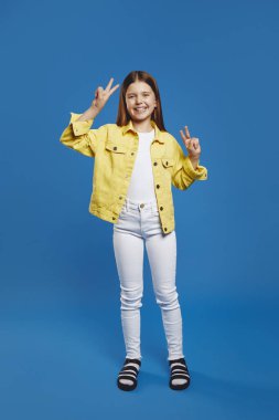 Gülümseyen çocuk sevinçle barış işaretleriyle poz veriyor, mavi bir zemine karşı canlı sarı bir ceket giyiyor. Gençlerin moda tanıtımları ve olumlu sosyal medya içeriği için ideal.