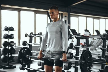 Uzun kollu atletik bluzlu ve şortlu bir adam modern bir spor salonunda halter kaldırıyor, antrenman sırasında odaklanmış duruyor. Fitness, antrenman, vücut geliştirme ve sağlıklı yaşam tarzı kavramları