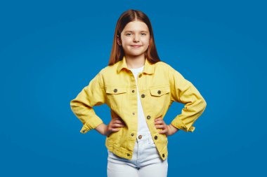Gülümseyen çocuk, elleri kalçalarının üzerinde duruyor, parlak sarı bir ceket giyiyor, pozitiflik yayıyor. Reklam ve gençlik odaklı markalaşma için ideal.