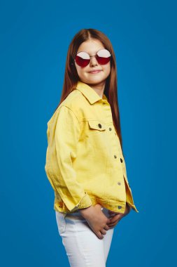 Parlak sarı bir ceket ve son moda güneş gözlüğü takan neşeli bir genç parlak mavi bir arka plana karşı duruyor, büyüleyici ve tarz dolu. Moda pazarlaması ya da çocuk giyim promosyonları için ideal..