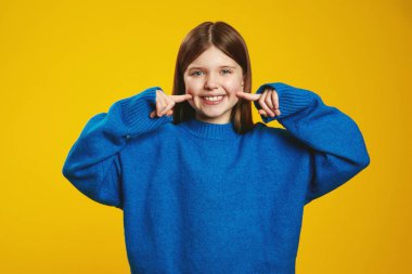 Parlak mavi süveterli mutlu çocuk yanaklarını işaret ediyor, parlak sarı bir arka plana pozitif enerji yayıyor. Reklam ve marka kampanyaları için ideal..