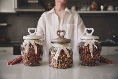 Kalp şeklinde kapaklı üç cam kavanoz bir mutfak tezgahında zarif bir şekilde yerleştirilmiş çeşitli fındıklar içeriyor. Beyaz gömlekli bir adam kavanozların arkasında duruyor ve sade bir atmosfer yaratıyor..