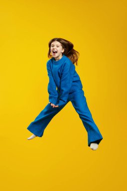 Neşeli bir gencin parlak mavi bir elbiseyle mutlu bir şekilde sıçradığı, canlı sarı bir arka plana neşe ve neşe saçtığı dikey bir fotoğraf. Çocuklar için ideal moda promosyonları ve mutlu yaşam tarzı
