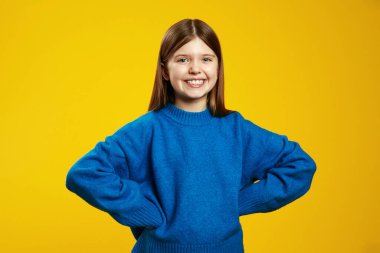 Rahat mavi bir süveter giyen neşeli çocuk, elleri kalçasında, parlak sarı bir arka plana karşı parlak bir şekilde gülümsüyor. Neşeli reklam ve pazarlama kampanyaları için ideal..