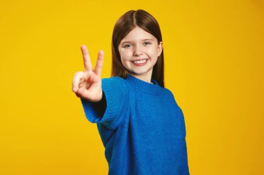 Gülümseyen çocuk, parlak sarı bir zemin önünde mutluluk ve pozitifliğin somutlaştığı bir barış işareti gösteriyor. Pazarlama, reklam ve sosyal medya içeriği için ideal.