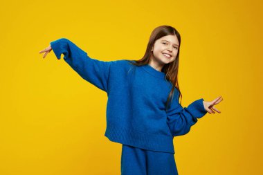 Mavi kazaklı gülümseyen çocuk, mutluluk ve enerji gösteren canlı sarı bir arkaplan karşısında sevinçle poz veriyor. Çocuk giyim reklamları ve yaratıcı pazarlama için ideal..