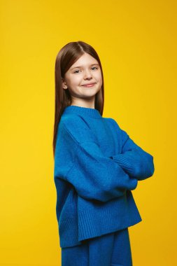 Mavi bir süveter giyerken içten bir şekilde gülümseyen neşeli ve kendinden emin bir çocuğun dikey fotoğrafı, canlı sarı bir arkaplan karşısında duruyor. Reklamlarda ve çocuk kampanyalarında kullanmak için ideal.