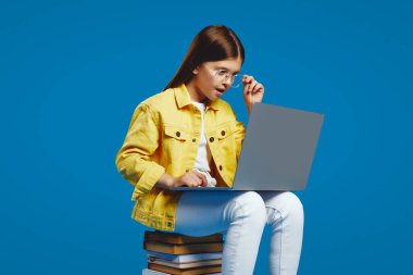 Sarı bir ceket giyen meraklı çocuk, kitapların üzerinde otururken dizüstü bilgisayarıyla oldukça meşgul, merak ve odaklanma gösteriyor. Eğitim içeriği ve teknoloji promosyonları için ideal.