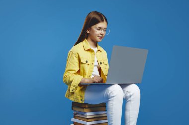 Çekici küçük kız dikkatle dizüstü bilgisayara bakıyor, bir yığın kitabın üzerinde oturuyor, konsantrasyon ve öğrenme yayıyor. Eğitim içeriği ve teknik promosyonlar için ideal.