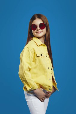 Parlak sarı ceket giyen sevimli bir çocuk ve modaya uygun kırmızı güneş gözlüğü takıyor, kendine güvenen bir tarzda parlak mavi arka plana doğru yayılıyor. Moda reklamları ve markalar için ideal..
