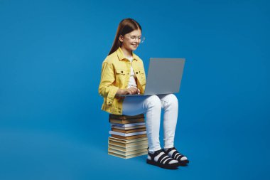 Yakışıklı, teknoloji meraklısı, gözlüklü, sarı ceketli ve beyaz pantolonlu bir kız uzun bir kitap yığınının üzerinde otururken dizüstü bilgisayarla çalışıyor. Parlak mavi arkaplan modern atmosferi güçlendirir.