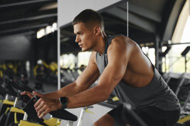 Modern jimnastik salonunda direksiyonu sıkıca tutarken ve ileriye bakarken konsantrasyonlu bisikletin yan görüntüsü. Spor ve sağlık hizmetleri kavramı.