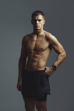 Kaslı erkek sporcu, bir stüdyoda üstsüz poz veriyor, atletik şortla güçlü ve formda. Sahne fiziksel dayanıklılığı ve aktif bir yaşam tarzına bağlılığı vurguluyor