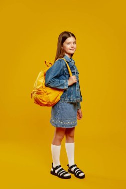 Kot giysili neşeli genç, parlak sarı bir sırt çantasıyla arka planda duruyor. Cazibesi ve kendine güveni tam. Okul promosyonları ve moda reklamları için ideal..