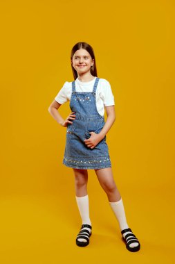 Kendine güvenen bir duruşu olan mutlu çocuk şık bir kot elbiseyle parlak sarı bir arka planda duruyor, gençlik cazibesini sergiliyor. Moda reklamları ve çocuk giyim promosyonları için ideal..