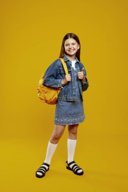 Sırt çantalı mutlu çocuk, parlak bir arka planda fotoğraf için poz verirken pozitifliğini ifade ediyor. Okul promosyonları ve pazarlama malzemeleri için ideal..