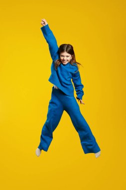 Mavi kıyafetli enerjik bir çocuğun dikey fotoğrafı büyük bir gülümsemeyle sevinçle sıçradı ve parlak sarı bir arkaplanda elini kaldırdı. Reklam ve yaratıcı pazarlama tasarımları için mükemmel..