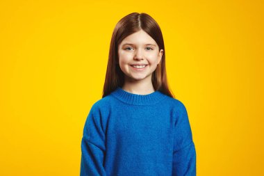 Mavi kazak giyen, uzun kahverengi saçlı mutlu genç bir çocuk, canlı sarı bir arkaplan önünde sıcacık gülümsüyor, pozitiflik ve neşe saçıyor. Çocuk giyim promosyonları için ideal.