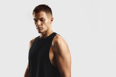 Siyah kolsuz bluz giyen kaslı bir gencin portresi, beyaz arka planda poz veriyor. Görüntü güçlü, atletizm ve zindelik taşır. Sağlıklı bir yaşam tarzı sergiler.