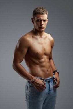 Kaslı ve kaslı bir adam, gri arka plana karşı izole edilmiş, kendinden emin, güçlü ve odaklı bir şekilde ayakta duruyor. Fitness markalaşma ve moda pazarlama materyali için ideal.