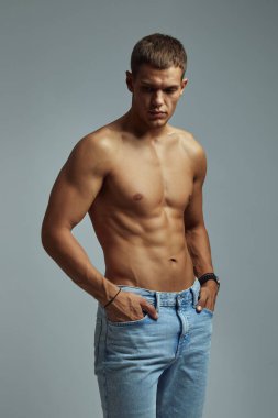 Atletik çocuk kot pantolon içinde üstsüz poz veriyor, gri stüdyo arka planına karşı kaslı ve renkli fiziğini sergiliyor, fitness, güç ve sağlıklı yaşam tarzını yansıtıyor..