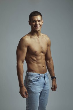 Ton gibi vücudu olan neşeli, üstsüz genç bir adam sıcacık gülümsüyor, günlük kıyafetlerle poz veriyor. Sağlıklı görünüşü ile fitness, pozitiflik ve özbakımı teşvik eder, gri arka planda yakalanır.