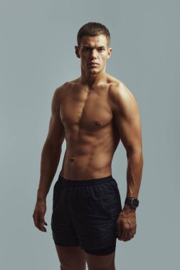 Spor şortu ve spor saati giyen kaslı genç bir adam, tarafsız bir stüdyo ortamında güven içinde poz veriyor, fiziksel uygunluğu, disiplini ve sağlık yaşam tarzını vurguluyor..