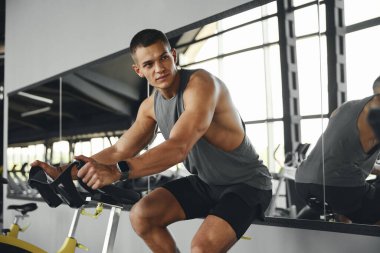 Sporcu adam spor salonunda bisiklete biniyor ve yan bakıyor. Kaslı insanlar bisiklet makinesi istasyonunda egzersiz yapıyor. Spor ve sağlık hizmetleri kavramı.