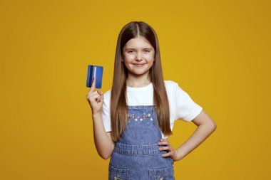 Neşeli genç bir kız, parlak sarı bir arkaplanın önünde kendine güvenen mavi bir kredi kartı sergiliyor. Finansal hizmetler veya çocuk pazarlama kampanyaları için ideal.