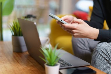 Masanın üstünde akıllı telefonu olan bir kadın emlak salonunun içinde sarı kanepe yeşil bitkiler tutuyor.