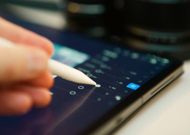 Dijital stylus tablet ekranı kullanan yakın plan kişi görüntü parametrelerini ayarlar, muhtemelen Lightroom. Arayüz kaydırıcıların pozlama, kontrastlama ve röfle düzenlemelerini gösteriyor. Modern dijital iş akışı, yaratıcı süreç, fotoğraf düzenleme, mobil reto