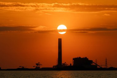 Endüstriyel bir fabrikanın arkasında gün batımı ve önünde deniz, turuncu renk, siluet