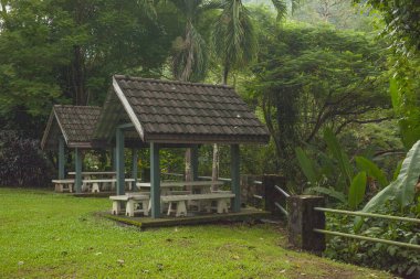 Parktaki iki pavyonun beyaz masaları ve sandalyeleri var. Arka planda yeşil ağaçların arasında metal bir çit..