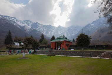 Public park in Srinagar There are two buildings. One has a red entrance and a green roof. A walkway and a large mountain range in the background
