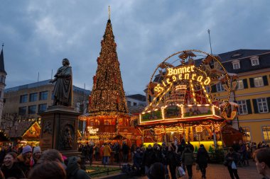 Bonn, Almanya - 6 Aralık 2023: Şenlik ışıkları, bir atlıkarınca ve Ludwig van Beethoven heykelinin arkasında büyük süslü bir Noel ağacı bulunan hareketli bir Noel pazarının alacakaranlık görüntüsü