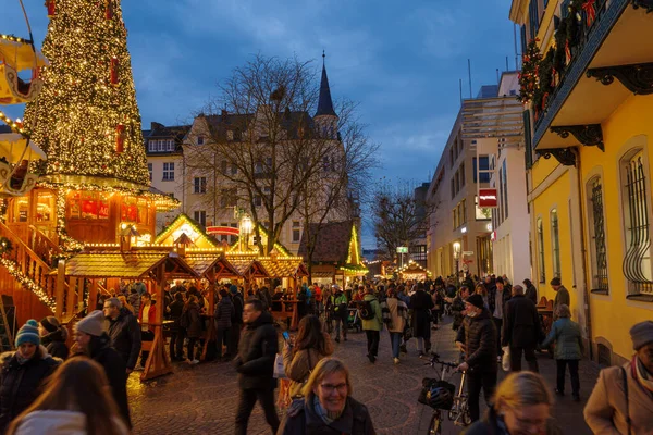 Bonn, Almanya - 6 Aralık 2023: Kalabalık bir Noel pazarının manzarası şenlikli ışıklar, dekore edilmiş ağaçlar ve tatil tezgahları, uzakta büyük ışıklı bir acil çıkış işareti ile.