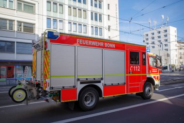 Feuerwehr Bonn 'dan gelen kırmızı itfaiye aracı arka planda binalar ve boş yolda yanıp sönen ışıklar olan bir şehir caddesinde hareket halinde..