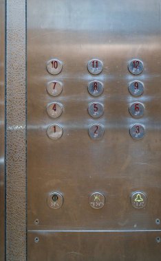 12 katlı asansör kontrol panelinin yuvarlak metalik düğmeleri aşınma izleri taşıyor..
