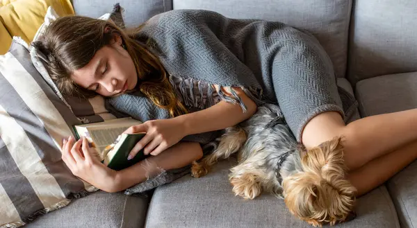 Genç kız modern bir kanepede uzanıp kitap okuyor. Uluslararası kitap günü. 23 Nisan. Gençlerde kitap okumanın önemi. Kitap günü reklamları ve kampanyaları için yatay fotoğraf..