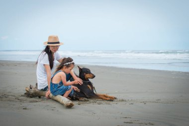 Anne, kız ve sahilde oturan bir köpek. 