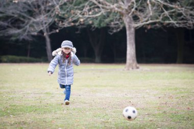 Küçük kız parkta yeşil çimlerde futbol topuyla oynuyor.