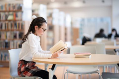 Asyalı üniversite öğrencisi kütüphanede kitap okuyor.