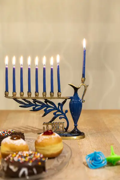 Menorah (Chanukkia) with 8 lit burning candles for Jewish Hanukkah holiday on table at home. Celebrating Chanukah festival of lights. Dreidel and Sufganiyot donuts sweet cultural food on a plate.