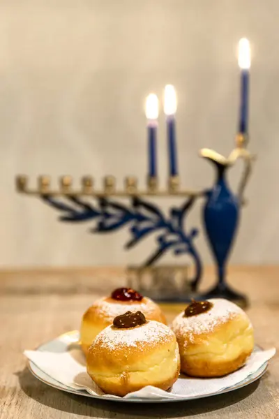 Menorah with lit burning candles for Jewish Hanukkah holiday on table at home. Celebrating Chanukah festival of lights.  Sufganiyot donuts sweet cultural food on a plate