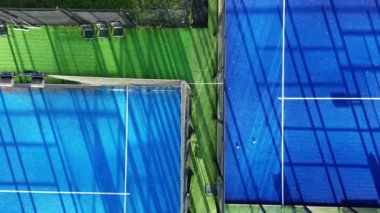 Mavi yüzey boyunca uzanan uzun gölgeli iki tenis kortunun havadan görünüşü. Mahkemeler yeşil çimlerle çevrili..